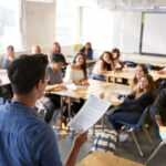 Rear View Of Male High School Teacher Standing At Front Of Class Teaching Lesson
