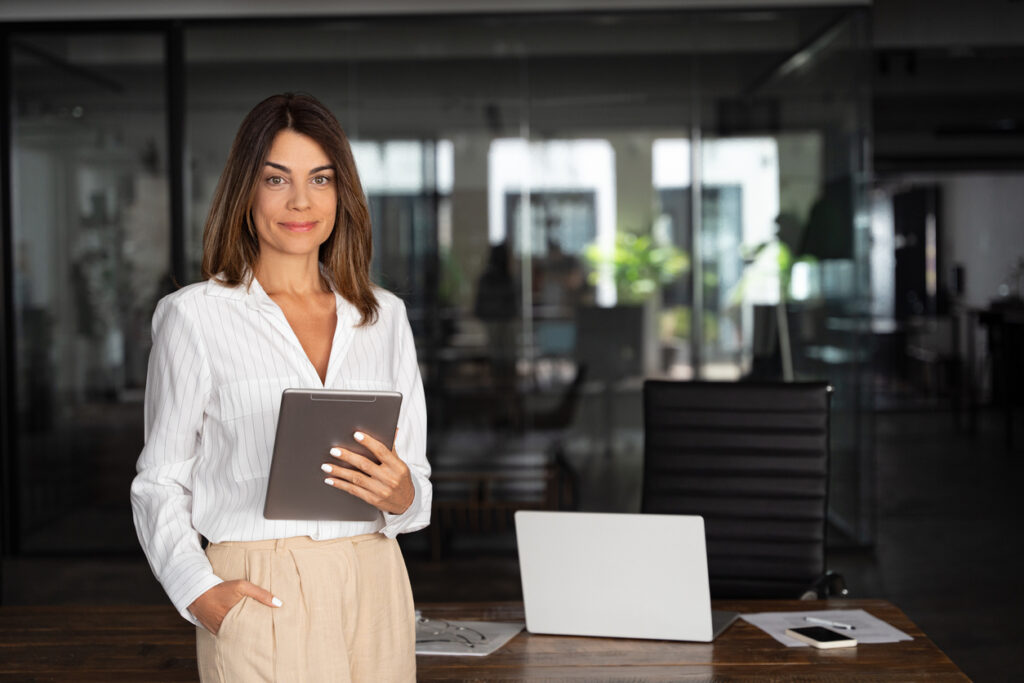 Latin Hispanic mature adult professional business woman looking at camera and smiling. European businesswoman CEO holding digital tablet using fintech tab application standing at workplace in office.
